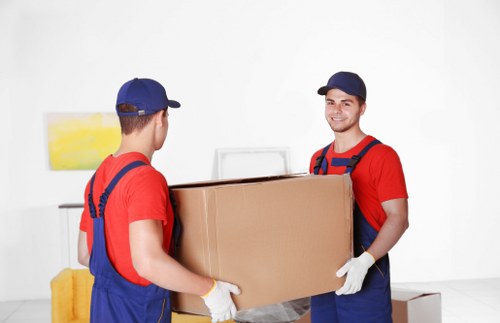 Two removal men with light skin and wearing red shirts, blue overalls, caps, and white gloves are in a clean, well-lit interior space, engaged in lifting and passing a large cardboard box together. One man faces away from the camera, holding the box from the left side, while the other, smiling and looking towards the camera, supports the box from the right. The background features a white wall with a small, colorful abstract painting on the left, and a yellow cushion or piece of furniture slightly visible behind the men. The scene appears to be in a residential setting, possibly a living room or hallway, with minimal furnishings in view. The box is standard brown, and the men's attire and positioning suggest they are providing removals or house moving services, consistent with Kensington Man with Van's offerings in the Kensington area of London, inferred from the page details. The environment is bright and tidy, emphasizing professionalism and careful handling of household items during a house relocation or removal process.