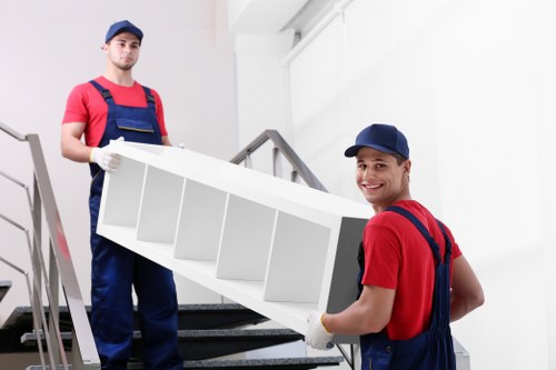 Two professional movers from Kensington Man with Van, dressed in red shirts, navy overalls, and blue caps, are carrying a large, white, multi-compartment shelving unit. One mover is positioned on a staircase, gripping the bottom of the unit with both hands, while the other stands on the step above, supporting the top. The shelving unit appears to be made of painted wood or similar material, with evenly spaced compartments suitable for household storage. The scene takes place inside a modern residential building with white walls, where natural light illuminates the space, reflecting a clean and professional environment typical of house removals services in Kensington. Behind and around the movers, a boiler or radiator is visible on the wall, and the staircase features metallic handrails. The precise handling and positioning of household items, combined with the appropriate packing and lifting equipment, highlight careful moving techniques used by Kensington Man with Van for safe and efficient relocations within London postal areas such as the W8 postcode district.
