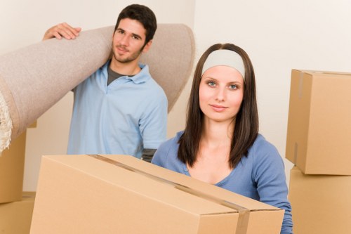 A young man and woman with light skin are inside a residential space, preparing for a house removal. The man, standing behind and to the left, has dark short hair and is wearing a light blue polo shirt, and is holding a large, light-colored upholstered sofa or armchair over his shoulder. He looks towards the camera with a neutral expression. The woman, positioned in the foreground to the right, has long dark hair and is wearing a white headband and a light blue top, holding a large cardboard box in front of her. The background shows a plain beige wall, and additional cardboard boxes are visible around them, indicating an organized packing or moving process. The environment appears well-lit with soft natural or artificial light, highlighting the textures of the furniture and boxes, with the setting suggesting an indoor hallway or living room space in a home in or near Kensington, where local house removal services are often provided by Kensington Man with Van.