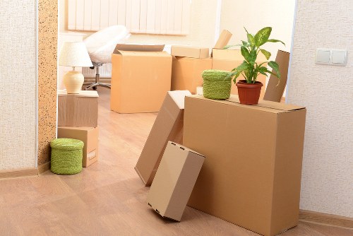 A room with light-colored wooden flooring and neutral wallpapered walls, showing several large cardboard moving boxes in various sizes stacked and placed along the space. On top of one of the larger boxes, there is a green potted plant with broad leaves, adding a touch of greenery to the scene. To the left, a small green woven storage basket is positioned on the floor. Towards the background, a white table lamp with a fabric shade sits on a light wooden side table, and a window above the table provides natural light. The room appears to be in the process of packing or moving, with some boxes open or closed, and items like a flat-screen TV box leaning against the large boxes, indicating an ongoing household move. The scene is framed as part of a house removal service operation, typical of Kensington Man with Van’s work in London neighborhoods near Kensington, with the focus on household contents and relocation activities within a residential interior.