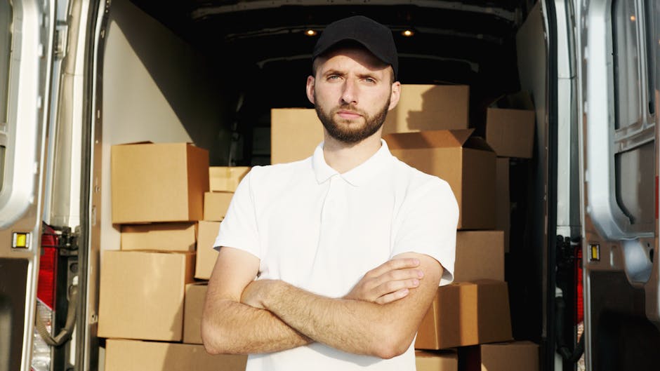 A man sitting on the edge of a large, open black van parked outside a residential property during daytime, surrounded by cardboard moving boxes of various sizes, some wrapped in plastic for protection