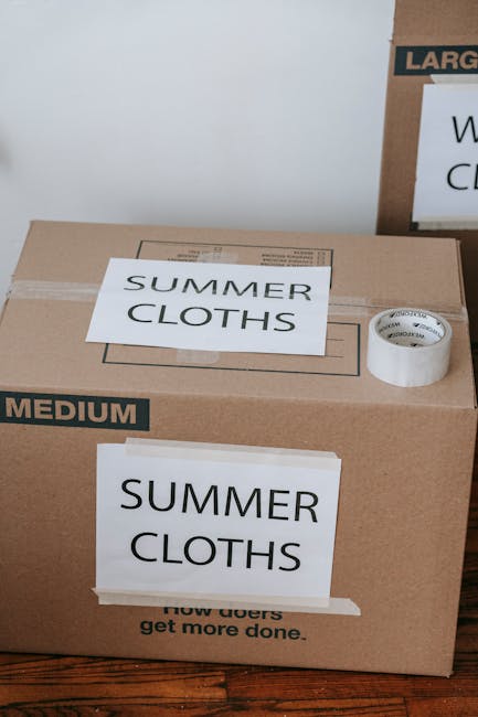 A young woman with long dark hair, wearing a white sleeveless top, beige pants, and striped sneakers, is sitting cross-legged on a wooden floor inside a room surrounded by large cardboard moving boxes
