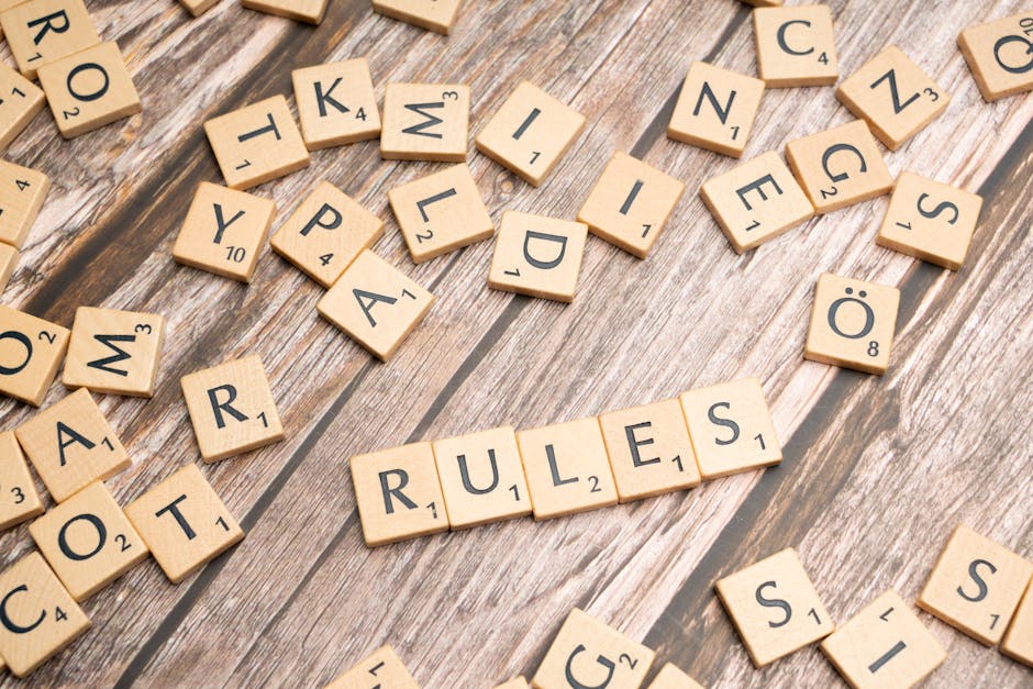 Close-up image of numerous Scrabble tiles scattered on a wooden surface, with a prominent set spelling out 'RULES' in the center. The tiles are made of light-colored wood with black letter and number