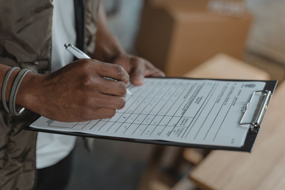Close-up image of a person's hand holding a pen and filling out a relocation or moving checklist on a clipboard, with a focus on the paperwork. The person is wearing a wristband, and the background sh